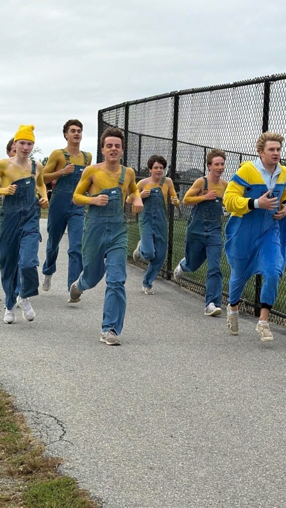 Homecoming 5K participants in yellow shirts and overalls running beside a chain-link fence.